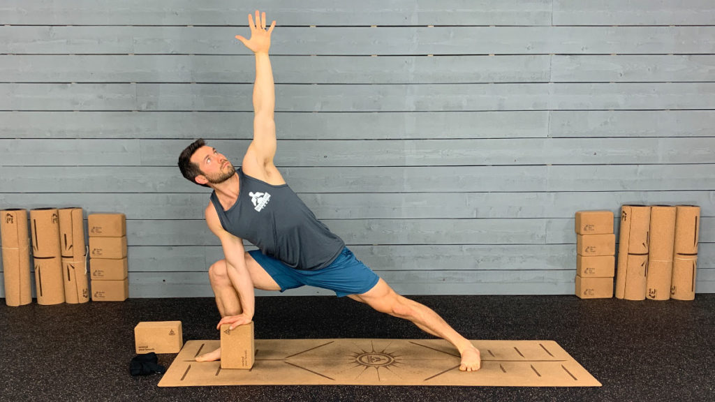 Yoga instructor demonstrates side angle with arm extended overhead pose using block on yoga mat