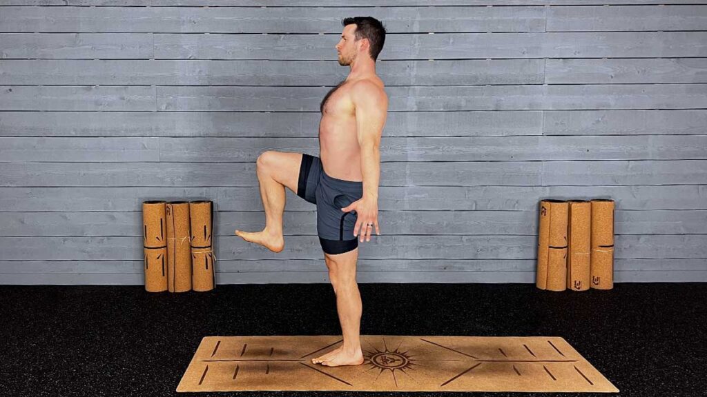Shirtless male yoga instructor demonstrates balancing on one foot standing march hold pose
